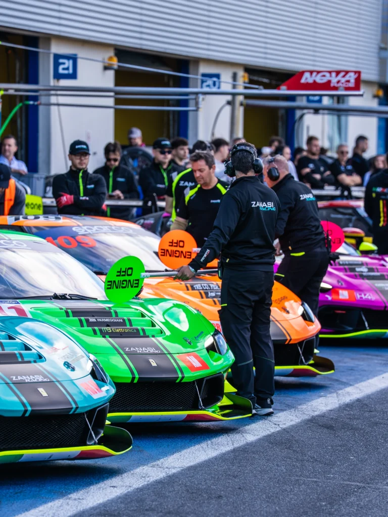 Crew members stand by a row of colorful race cars in a pit lane, with one holding a green Engine Off sign