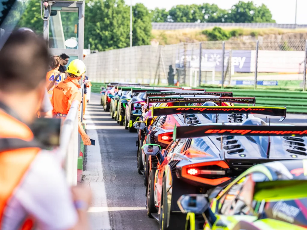 A line of colorful GT race cars queued up in a pit lane at a racetrack with a track marshal standing nearby