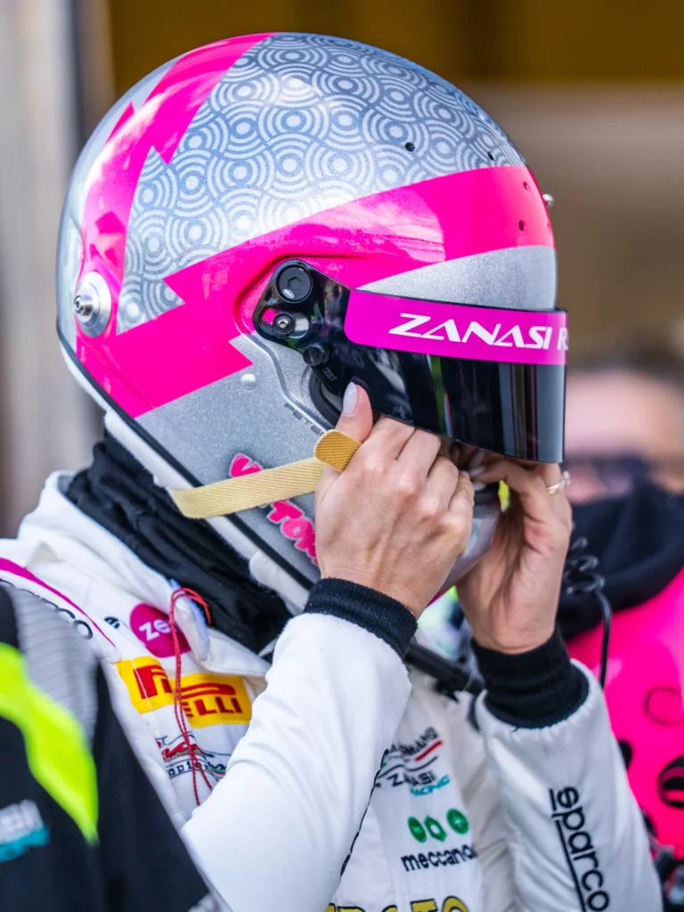 A racing driver adjusts a silver and pink helmet with a Zanasi visor