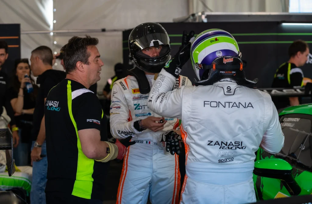 Two Zanasi Racing drivers in white suits and helmets talk with a crew member in a garage