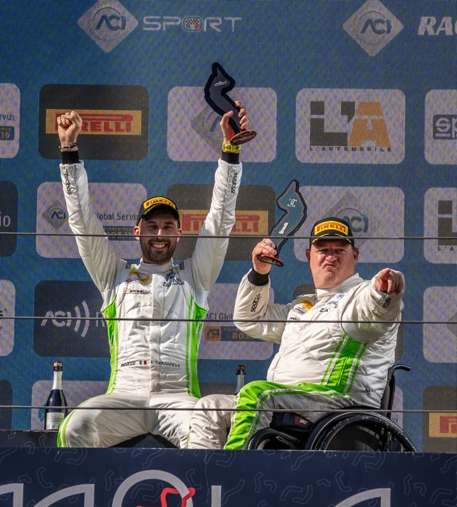 Two racing drivers celebrate on a podium with trophies; one driver is in a wheelchair